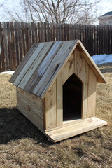 wooden doghouse on dry grass with arched entrance and light planks in rustic backyard setting with early spring atmosphere