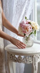 mediterranean coastal baroque light vintage bedroom, white carved wood side table near open window, lace curtains, woman's hands placing vase