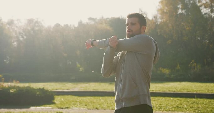 Close view of Caucasian male with beard and stylish haircut stretching upper body in middle of park. Sun shining very bright in background. Exercising in morning before work. Caring about health.