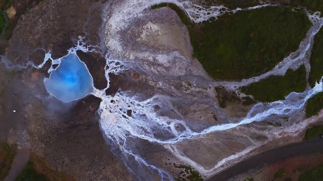 Aerial top down view of a turquoise hot spring in Haukadalur, Iceland, white mineral deposits, braided runoff to a road, thin steam, high altitude, saturated contrast