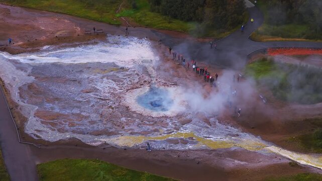 Aerial top down view shows blue vent, steam, sulfur yellow streaks, and sinter terraces, with tourists on paths at Haukadalur, Iceland, under overcast daylight.
