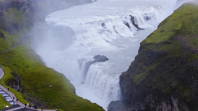 Aerial shot of Gullfoss on the Hvita river shows multi tiered falls plunging into a rocky canyon, mist rising, visitors on paved paths, vivid green moss, high contrast look.