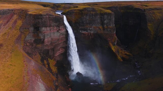Aerial drone shot shows Haifoss plunging into a basalt canyon in Iceland. A rainbow forms in mist, river threads a narrow gorge, power lines mark horizon, cool diffuse light.