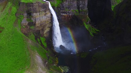 Aerial view shows Haifoss plunging over columnar basalt into a narrow canyon in Iceland, with a rainbow in mist, mossy cliffs, summer greens, and deep shadows. - Powered by Adobe