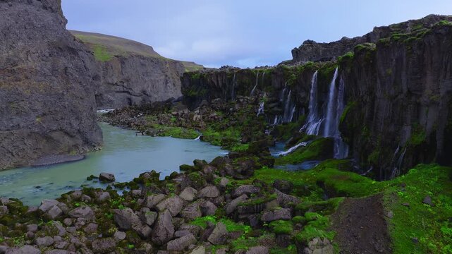 Aerial view shows Sigoldugljufur canyon in Iceland, dark basalt cliffs with multiple slender waterfalls feeding a milky turquoise glacial river, mossy rocks, soft overcast light