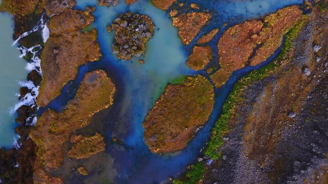 Top down aerial shows milky turquoise channels through mossy islands and dark basalt, foaming surf and a small cascade near Vatnajokull in autumn light.