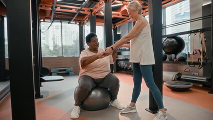 Full shot of mature Caucasian woman in sportswear helping her plus size mature African American female friend to maintain balance while sitting on fitness ball, exercising together in modern gym - Powered by Adobe