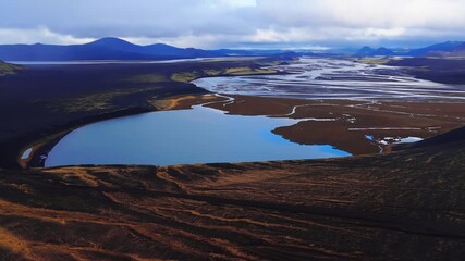 Aerial view shows cobalt lagoon feeding braided meltwater channels across black sand plains, with ochre ridges and volcanic cones under diffuse daylight in Iceland. - Powered by Adobe