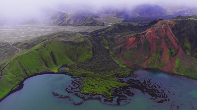 Aerial view in Landmannalaugar, Iceland shows rhyolite mountains, lava islets, a turquoise lagoon, braided river and sandur as the camera glides in soft overcast light.