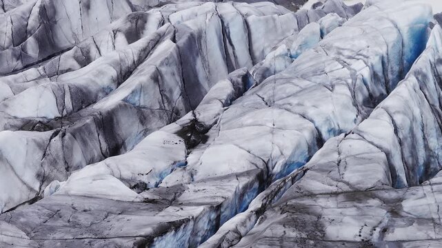 Aerial oblique view shows a crevassed glacier tongue in Iceland with electric blue ice, dark volcanic ash streaks, and pale meltwater pools under cool overcast light.