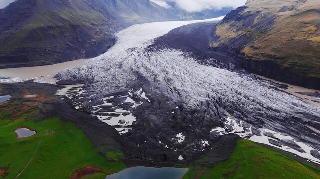 Aerial view shows an ash darkened glacier tongue in southeast Iceland meeting braided, milky rivers and ponds, with a heart shaped tarn, mossy meadows, and overcast light.