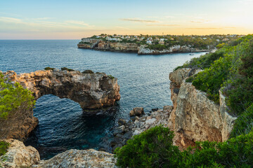 Obraz premium Majestic stone arch in sea at Cala Santany?, Mallorca, Spain during sunset. Serene seascape with glowing sky and coastal rock formations.