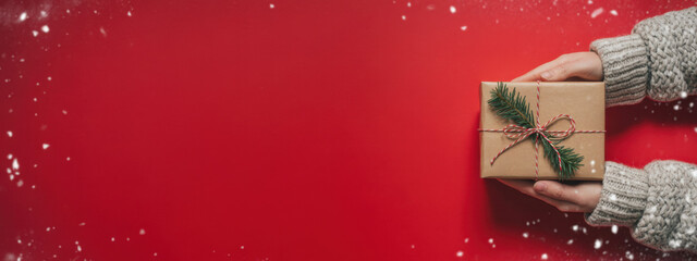 Female's hands in pullover holding Christmas gift box decorated with evergreen branch on red background with snow. Christmas and New Year banner.
