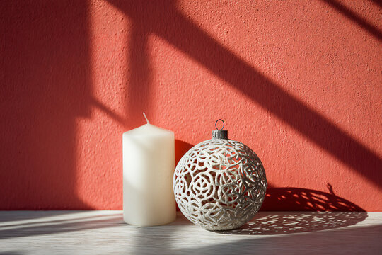 White pillar candle and silver filigree Christmas ornament with strong sunlight shadow on red textured wall, minimalist festive still life, modern holiday decor photography