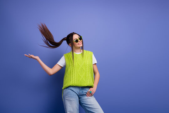 Young woman in bright lime green knit vest and white tee flips her hair with a playful smile against a blue studio background