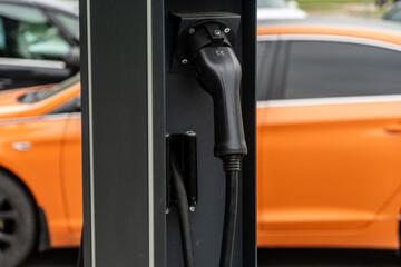 Electric vehicle charging station with charging cable and orange car in background during daytime
