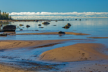 Rocks dot shallow lake water near sand beach with cloud lined distant horizon.