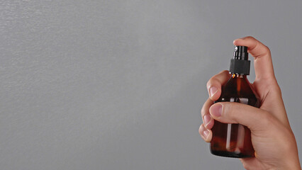 Close-up of a female hand spraying a fine mist from a amber bottle on a grey background. Skincare or haircare spray serum in use with copy space. Spraying process wellness cosmetic beauty product 