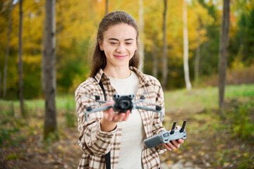 A person is enjoying outdoor drone flying in a colorful forest. They are smiling while holding the drone in one hand and the controller in another, surrounded by autumn trees.
