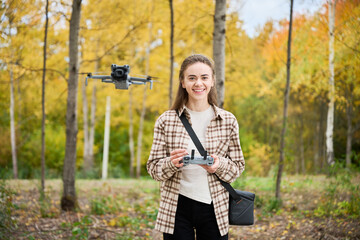 In a lively forest adorned with autumn colors, a young woman smiles as she skillfully pilots a drone. The trees around her showcase orange, yellow, and green hues.