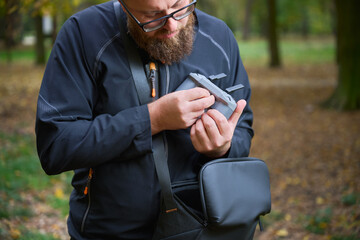 A person examines a camera accessory closely while standing in a beautiful autumn park. Surrounding yellow and orange leaves create a peaceful atmosphere.