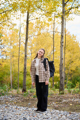 A woman stands happily in a forest surrounded by golden autumn leaves. She carries a backpack and smiles warmly at the camera, showcasing the beauty of nature.