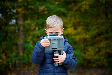 A boy dressed in a blue jacket holds a camera, focused and curious, surrounded by vibrant autumn leaves and trees. The scene captures childhood wonder with nature.
