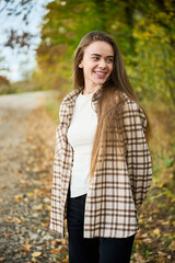 A young woman with long hair stands beside a gravel path, smiling happily as the autumn leaves surround her. The sun shines through the trees, creating a warm glow.