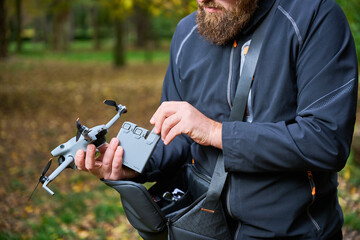 A man with a bushy beard prepares his drone for flight in a peaceful park filled with trees. He carefully adjusts the device on a calm, sunny day.