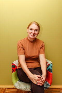 Smiling middle-aged woman with red hair in brown top sits on colorful patchwork chair against light green wall. portrait of a middle-aged woman 45 years old, with a warm smiling expression