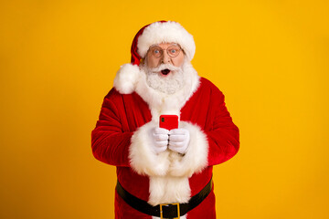 Santa in red suit with white beard holds a smartphone against a bright yellow background for festive holiday magic and shopping vibes