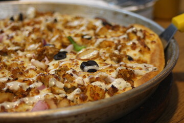 Close-up view of a deep-dish pizza, featuring large chunks of seasoned chicken, red onions, black olives, and a drizzle of creamy white sauce, sitting in a metal pan.