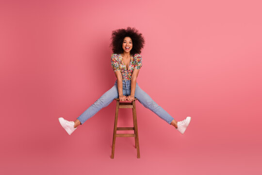 Cheerful young woman with curly hair sitting on a wooden stool on a vibrant pink studio background showcasing joyful vibes - Powered by Adobe