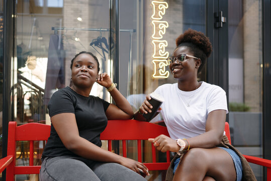 Two young women sitting outside a coffee shop with smartphones and takeaway coffee, talking and laughing, representing friendship, social connection, and modern city lifestyle.