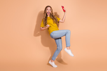 Excited young woman in casual outfit holding phone and smiling on beige background