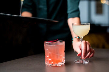 A bartender serves two cocktails, a pale yellow drink in a coupe glass and a pink drink in a faceted rocks glass garnished with a rose petal, on a dark bar counter