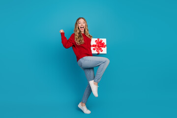 Cheerful young woman in red sweater holding a gift with ribbon against a blue background showcasing...