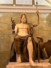 Colossal marble statue of the seated god Zeus (Jupiter) holding a small figure of Nike, with an eagle beside him, set against an ornate green marble wall © AndreyZayats