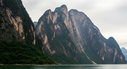 Majestic towering green mountains rise dramatically from a calm reflective body of water under a cloudy sky