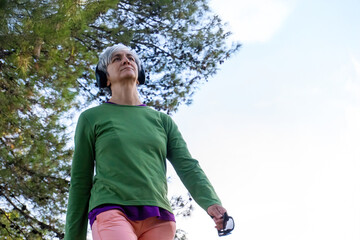 Mature woman with short gray hair and headphones walking outdoors through a green park, enjoying physical activity and nature on a sunny morning.
