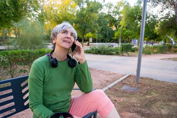 Smiling mature woman with short gray hair sitting on a bench in a park, talking on her smartphone and enjoying a cheerful outdoor conversation.