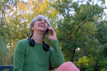 Happy woman with short gray hair sitting in a park and laughing while talking on her smartphone, enjoying a joyful outdoor conversation.