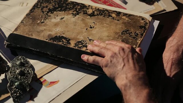Elderly hands examine an antique book with a crumbling cover, intricate metal jewelry, and a woven traditional textile. Preserving historical artifacts and craftsmanship research.