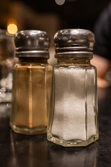 Glass salt and pepper shakers on dark restaurant table under warm evening lighting. Simple detail of everyday dining atmosphere.
