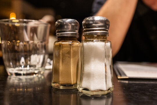 Glass salt and pepper shakers on dark restaurant table under warm evening lighting. Simple detail of everyday dining atmosphere.