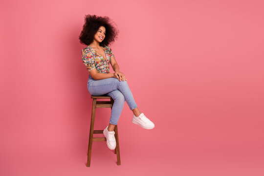 Positive woman with curly hair smiling confidently while sitting on a wooden stool against a vibrant pink background