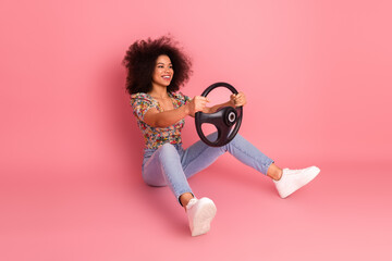 Cheerful young woman holding a steering wheel against a pink background exuding positivity, fun and...