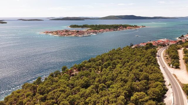 Stunning aerial view of a picturesque coastal road winding through lush green forests, leading to a charming island Krapanj town surrounded by the sparkling turquoise waters of the Adriatic Sea. 