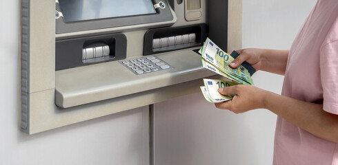 Woman withdrawing euro banknotes from an ATM, holding cash and bank card. Concept of euro currency, cash withdrawal, modern banking and financial transition in Europe. Horizontal banner