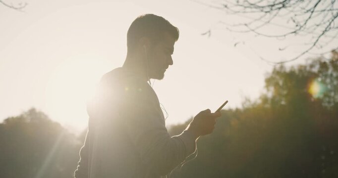 Gorgeous Caucasian male passing trees while checking video or playlist on smartphone. Typing message on mobile device. Beautiful sunset in background. Walking in part. Remote communication.